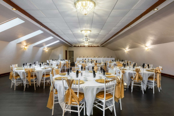 An elegantly decorated banquet hall set with round tables covered with white linens, golden accents, chairs with white covers and golden sashes, and chandeliers.