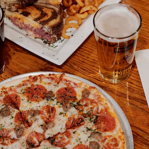 The image shows two drinks, a pepperoni pizza, curled fries, a sandwich with meat and cheese, and a burrito on white plates on a wooden table.