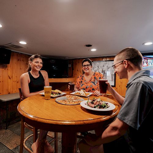 Three people are sitting at a round table in a wood-paneled room, enjoying food and drinks with a television in the background.