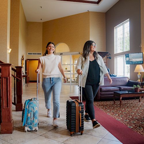 Two women are walking through a hotel lobby with their suitcases. One wears a white top, and the other a black jacket over a white shirt.