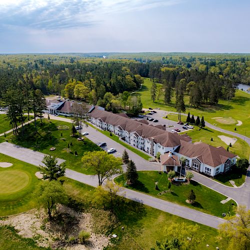 Aerial view of a large building complex surrounded by greenery, a golf course, and a river, with a forested area in the background.