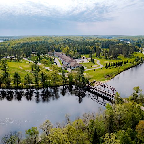 Aerial view of a lush green landscape with a river, buildings, a bridge, and extensive forested areas under a partly cloudy sky ending the sentence.