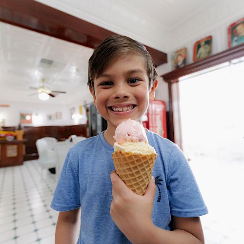 A young boy is smiling while holding an ice cream cone inside a brightly lit and retro-styled ice cream parlor, wearing a blue shirt.