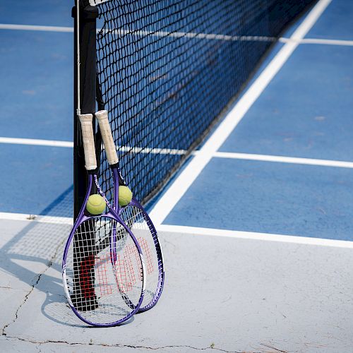The image shows two tennis rackets and two tennis balls leaning against a tennis net on a blue tennis court, with visible court lines.