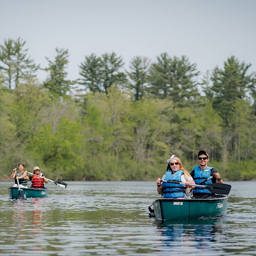 People are paddling canoes on a calm lake surrounded by trees, wearing life jackets and enjoying the outdoor activity.