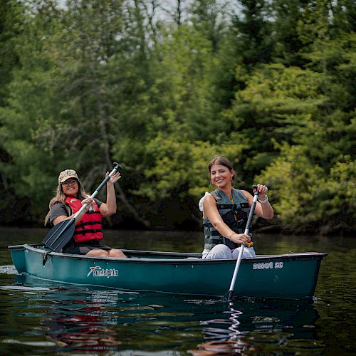 Two people are paddling a green canoe on a calm lake, surrounded by lush green trees, both wearing life vests and smiling.