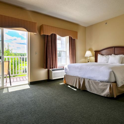 A hotel room with a large bed, a lamp, and a windowed door leading to a balcony with outdoor seating.