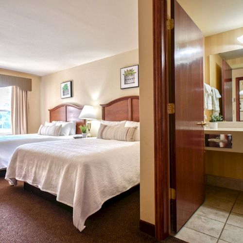 The image shows a hotel room with two beds, a TV, a window, and an adjacent bathroom with a sink and mirror.