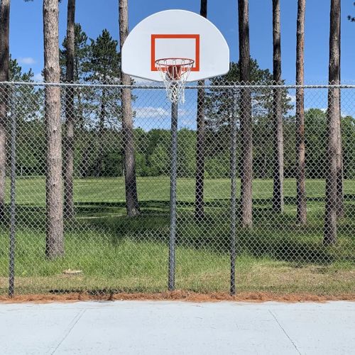 A basketball hoop stands in front of a chain-link fence, with tall trees and a grassy field in the background under a clear blue sky.