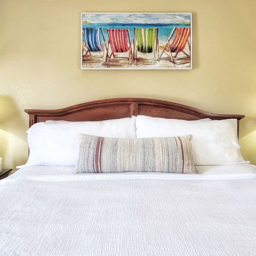 A neatly made bed with white linens, flanked by two lamps on side tables, and a beach chairs painting above the headboard.