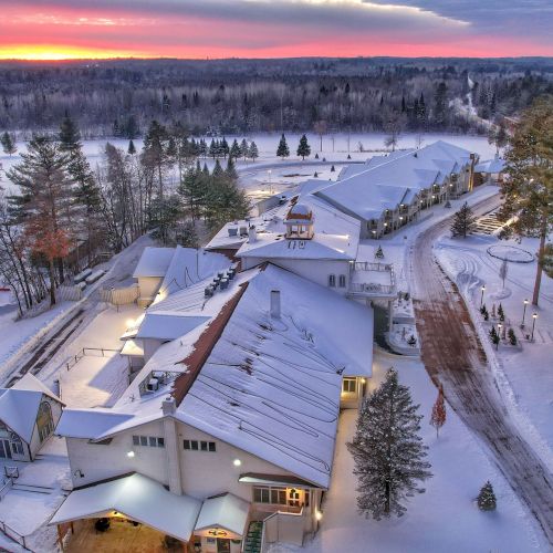 A snow-covered building complex surrounded by trees at sunset, with a winding path and a forested area in the background.