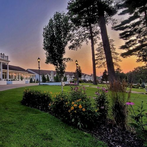 A large white building with a dome, surrounded by green lawns and colorful flowerbeds, set against a sunset sky with tall trees.
