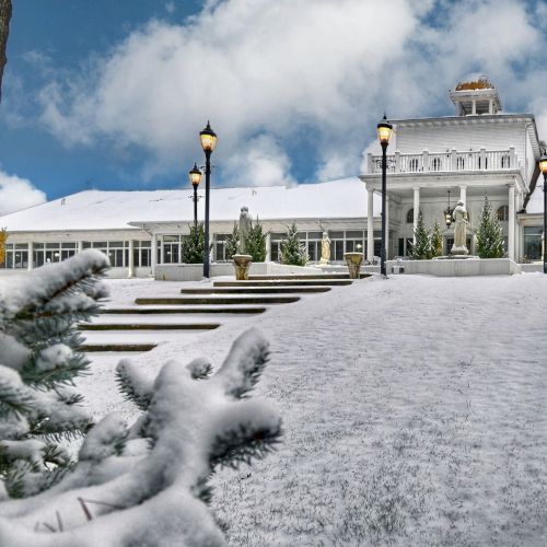 A snowy landscape with a large building, surrounded by lampposts and statues, with a cloudy sky in the background.