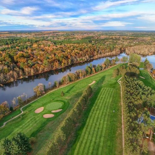 Aerial view of a golf course surrounded by trees, near a river with a backdrop of a vast, scenic landscape under a blue sky.