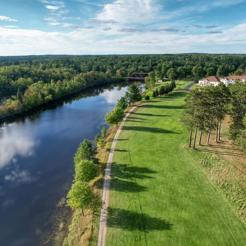 A scenic view of a river, a grassy area with trees, and a few houses in a lush, forested landscape under a partly cloudy sky.