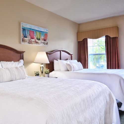 The image shows a hotel room with two beds, a nightstand with a lamp, a painting above the bed, and a window with curtains.