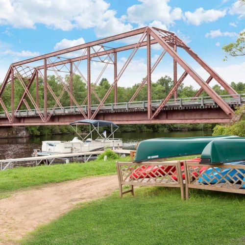 A metal bridge spans over a river with boats docked below; canoes rest on the grass nearby, surrounded by lush greenery.