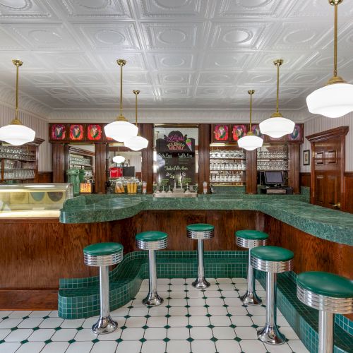 A vintage-style diner with green stools, retro lighting, and a classic counter setup, featuring wooden accents and tiled flooring.