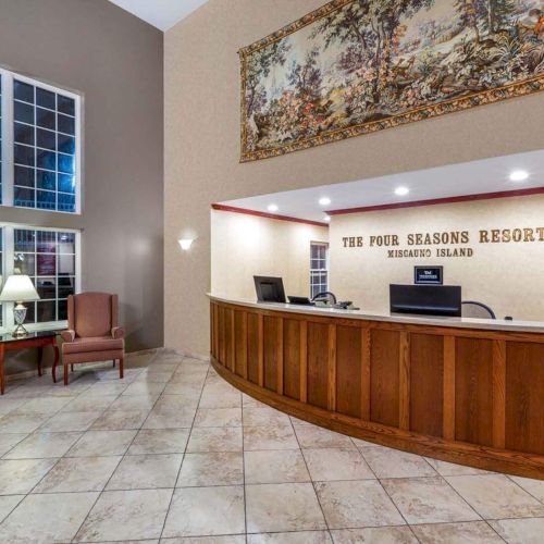 This image shows a hotel reception area with a curved wooden desk, armchairs, a lamp, large windows, and a decorative tapestry on the wall.