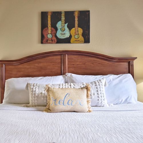 A neatly made bed with a "relax" pillow, flanked by lamps, and a painting of guitars on the wall above.