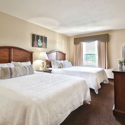 The image shows a hotel room with two double beds, a TV, a window, a lamp, and a framed picture on the wall.