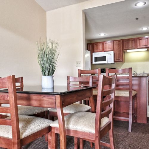 A dining area with a wooden table, four chairs, and a plant; visible adjacent kitchen with cabinets and appliances.