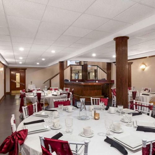 The image shows a banquet hall with round tables set for an event, featuring white tablecloths, red chairs, and a wood-paneled bar area.