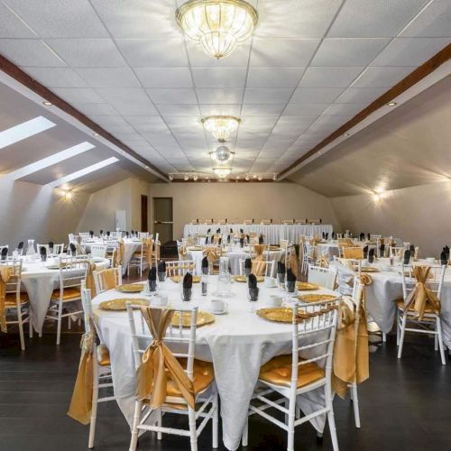 A banquet hall with round tables, white tablecloths, and golden accents is set up for an event.