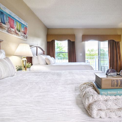A hotel room with two beds, a painting, a lamp, and books with glasses on one bed. There's a view of greenery through large windows.