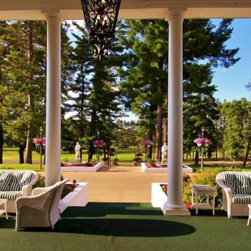 A scenic outdoor patio with white columns, comfy seating, and statues, overlooking a tree-filled landscape under a clear blue sky.
