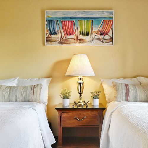 A cozy bedroom with two beds, a wooden nightstand, and a lamp, topped with a beach-themed painting on the wall.