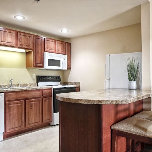 A modern kitchen with wooden cabinets, stove, microwave, fridge, dishwasher, and a small breakfast bar with chairs.