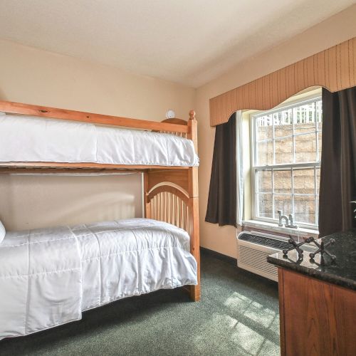 A room with wooden bunk beds, white bedding, a window with curtains, a lamp on a dresser, and a small plant decorates the surface neatly.