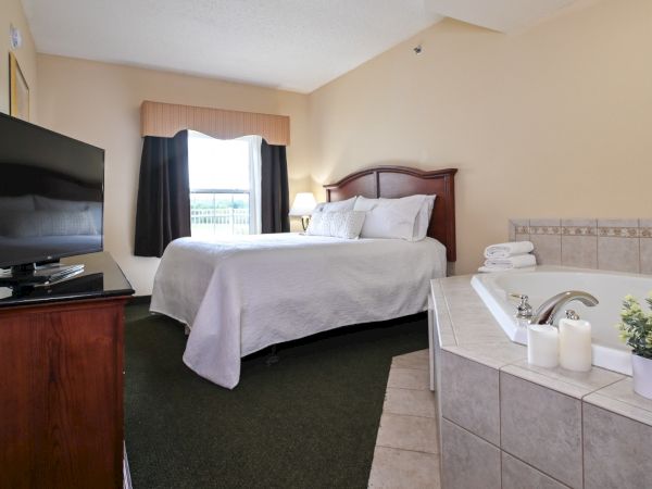 The image shows a hotel room with a bed, a flat-screen TV on a wooden stand, a window, a bedside lamp, and a bathtub with towels and toiletries.