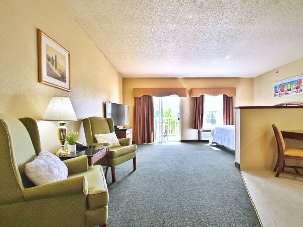 A cozy hotel room featuring two armchairs, a TV, a bed, a small dining area, and a balcony with an open sliding door allowing natural light in.