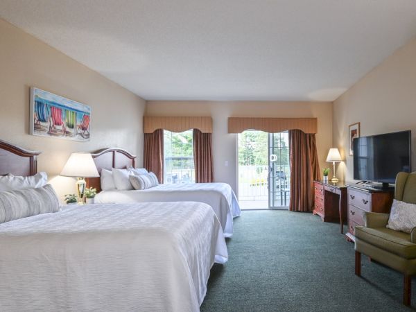 A hotel room with two beds, a TV, a large window with curtains, a green armchair, and a balcony outside.