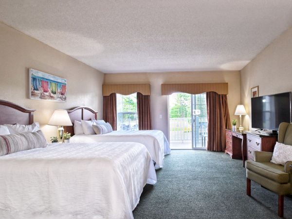 A spacious hotel room features two double beds, a flat-screen TV, an armchair, a desk, and large windows with a sliding door leading outside.