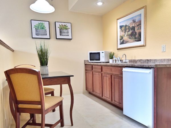 A kitchenette with wooden cabinets, a small white fridge, microwave, and a table with two chairs. Wall art and plants add decor.