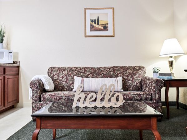 A cozy living room with a floral-patterned sofa, coffee table with "hello" decoration, and a table lamp. There's a framed painting on the wall.