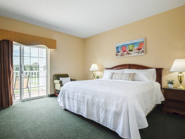 A well-lit bedroom features a large bed with white linens, two bedside tables with lamps, an armchair, and a painting, with sliding doors to a balcony.