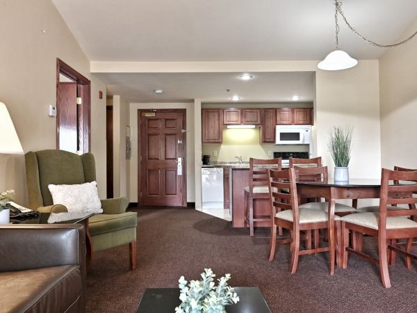 A cozy hotel room features a kitchenette, a dining table with chairs, an armchair, and a sofa. Decor includes a lamp and a potted plant.
