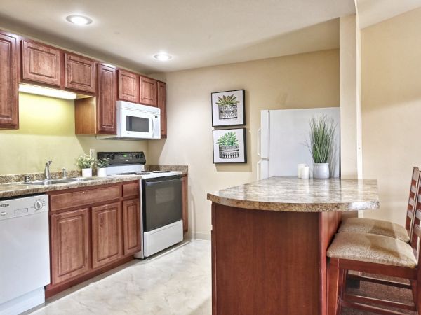 This image shows a small kitchen with wooden cabinets, a stove, microwave, dishwasher, refrigerator, and a breakfast bar with two chairs.