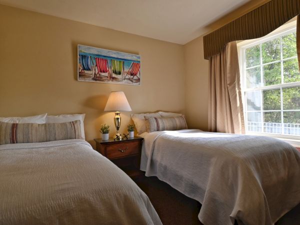 A cozy bedroom with two beds, a nightstand with a lamp, plants, and coastal-themed wall art. A large window lets in natural light.