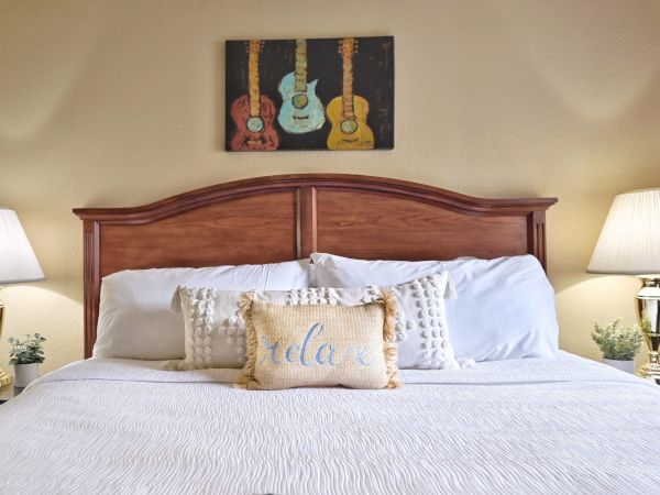 A neatly made bed with a "relax" pillow, flanked by lamps on nightstands, and a painting of guitars above the headboard.