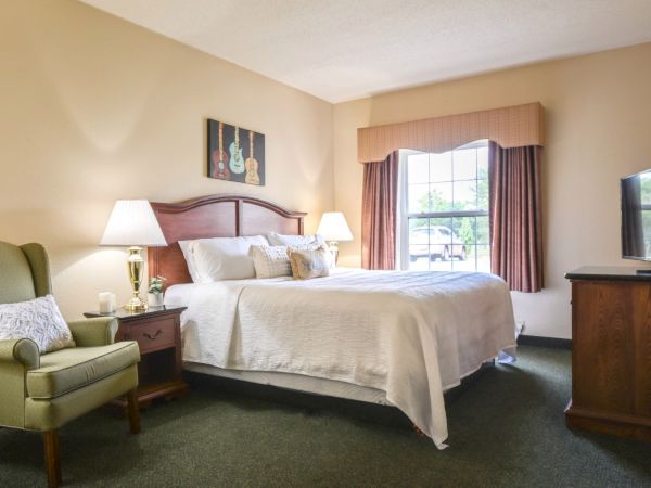A cozy bedroom features a bed, two nightstands with lamps, an armchair, a window with curtains, and a TV on a wooden dresser, creating a warm ambiance.