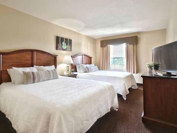 A hotel room with two beds, a TV on a dresser, a lamp between the beds, a window with curtains, and a guitar-themed painting on the wall.