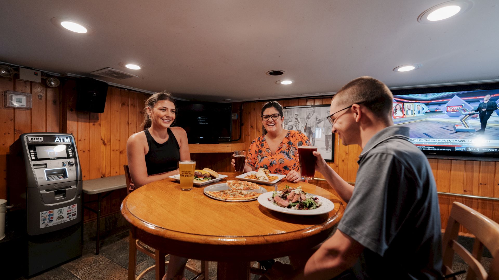 Three people are seated at a round wooden table, enjoying food and drinks in a cozy room with wood-paneled walls and an ATM.