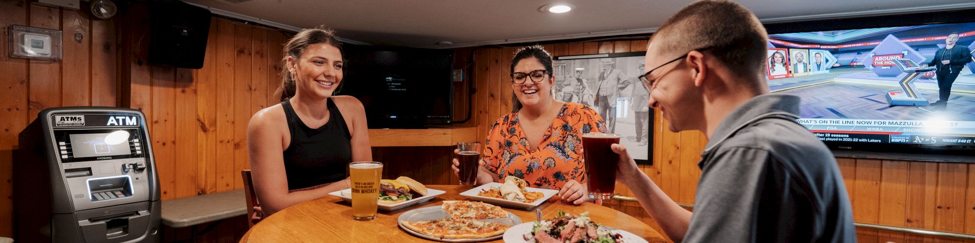 Three people are seated at a round wooden table, enjoying food and drinks in a cozy room with wood-paneled walls and an ATM.