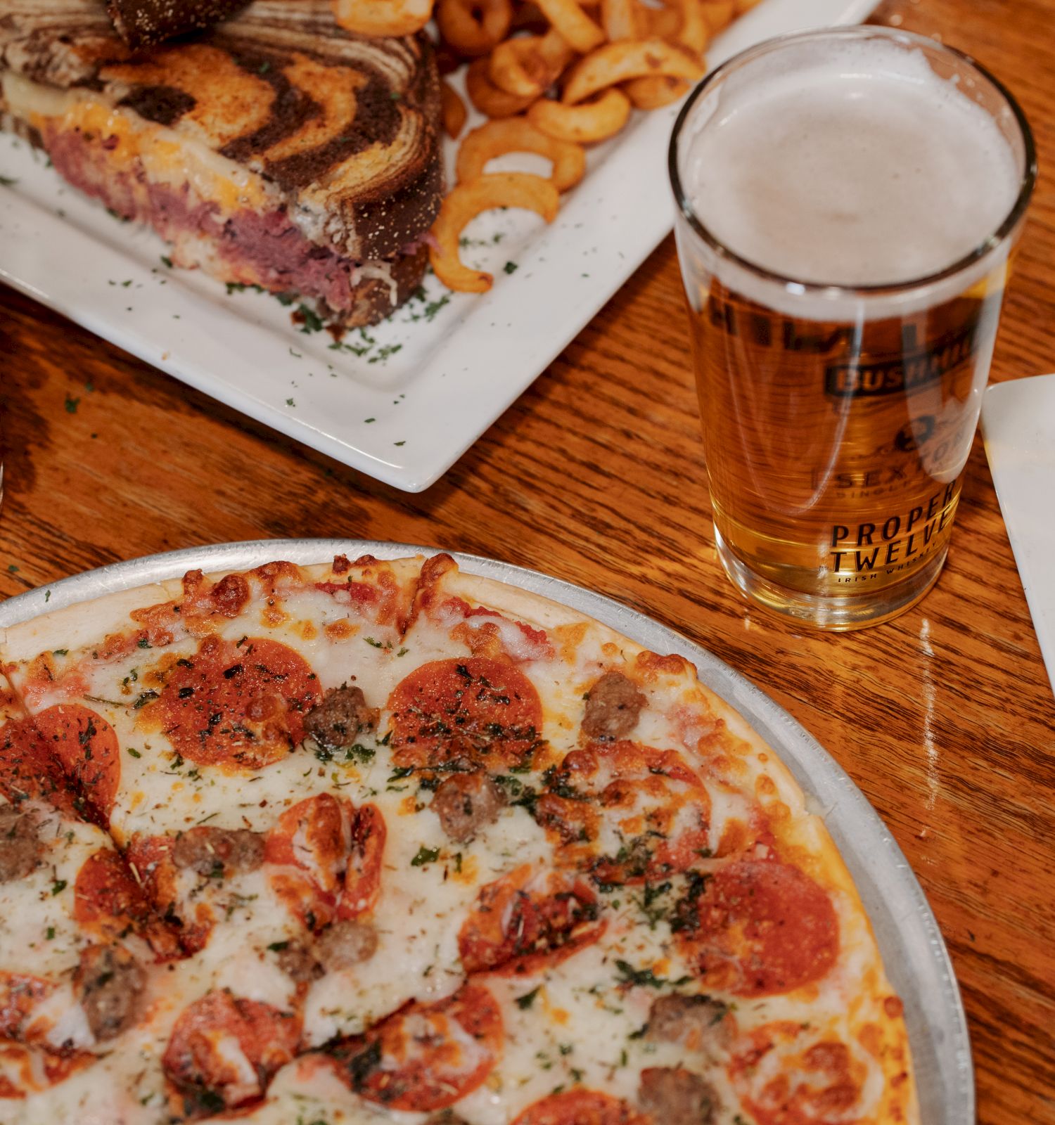 The image shows a table with a pizza, two glasses of beer, a sandwich with curly fries, and a plate with more curly fries and a burrito.