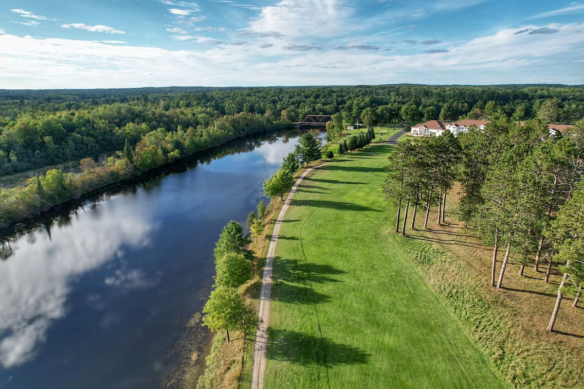 An aerial view shows a green park with trees and a lake alongside. Buildings can be seen in the distance against a backdrop of dense forests and a blue sky.